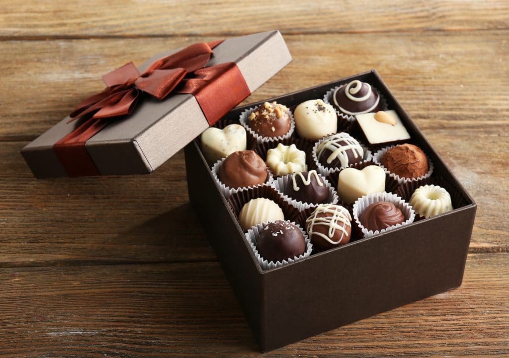 Hộp quà làm bằng bìa cứng với một chiếc nơ lớn màu đỏ.  Hộp được mở ra và bên trong là vài viên kẹo sô cô la với nhiều hương vị khác nhau.