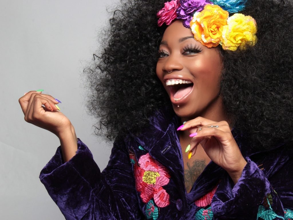 Hình ảnh một người phụ nữ da đen xinh đẹp.  Cô ấy rất vui và cười để lộ hàm răng trắng đẹp.  Cô mặc một chiếc áo cánh màu tím thêu hoa.  Tóc của cô cũng được trang trí bằng những bông hoa màu lớn.
