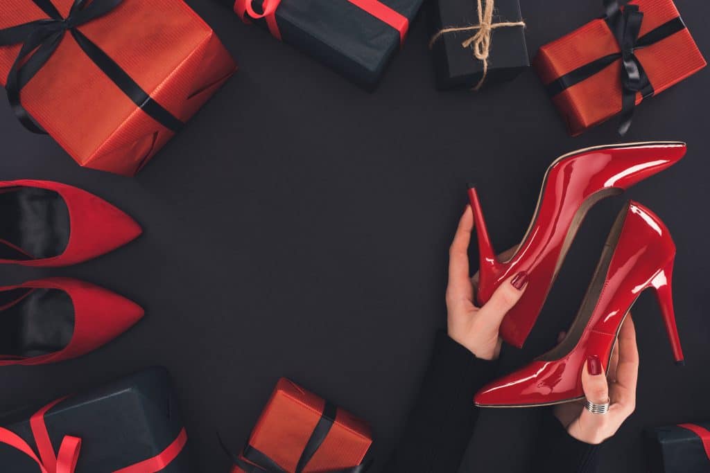 Hình ảnh một bàn tay phụ nữ với móng tay sơn đỏ.  Đôi tay này cầm một chiếc giày có gót bằng sáng chế màu đỏ.  Bên cạnh đó là một số hộp quà được gói bằng giấy đỏ và đen và trang trí bằng vải sa tanh màu đen và đỏ.