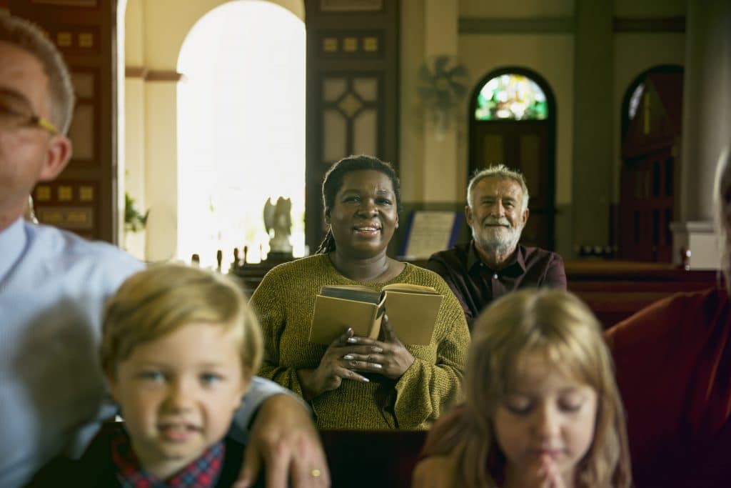 Người dân và trẻ em tham gia nghi lễ tôn giáo bên trong nhà thờ.  Một trong những người da đen và đang cầm cuốn kinh thánh.  Mọi người trong ảnh đều vui vẻ.