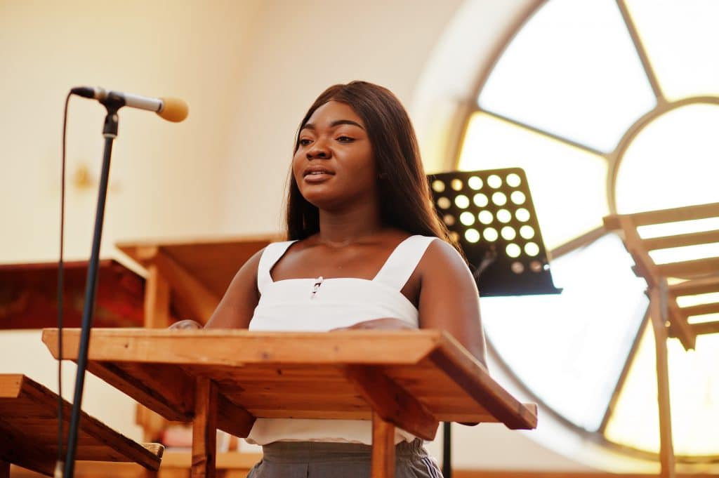 Người phụ nữ da đen có mái tóc dài, mặc áo blouse trắng.  Cô ấy đang ở bên trong nhà thờ, trước bục giảng và bên cạnh micro.  Cô ấy sẽ hát.