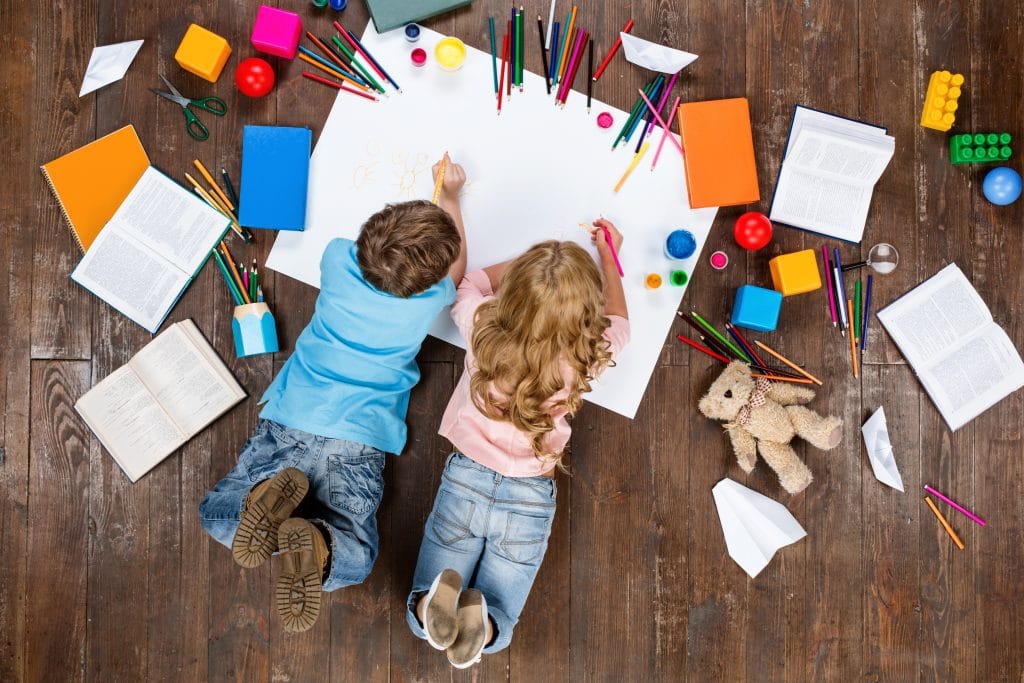 Hai đứa trẻ (một trai và một gái) nằm úp mặt trên một tấm thẻ trắng.  Họ đang vẽ và vẽ.  Xung quanh nó là vài cuốn sách, bút chì màu và đồ chơi nằm rải rác trên sàn gỗ tối.